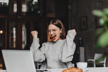 happy woman glasses makes winning gesture sincerely rejoices lady with red lipstick dressed gray sweater looking laptop
